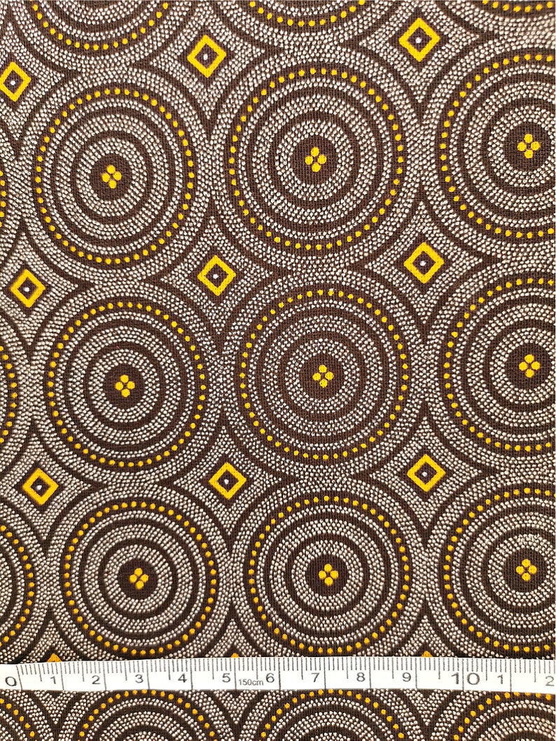 May include: Brown fabric with a repeating pattern of white and yellow circles and diamonds. The fabric is measured with a ruler in inches.