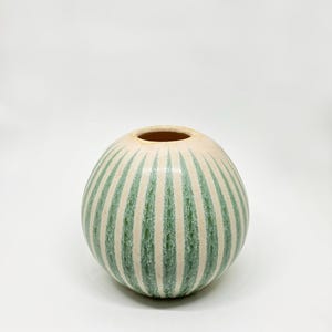 May include: A round ceramic vase with a cream-colored base and vertical green stripes. The vase has a wide opening and a smooth, glossy finish. The stripes are evenly spaced and add a decorative element to the vase.