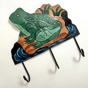 May include: A whimsical metal wall hook featuring a green frog design. The frog is painted with details and sits on a brown and orange background with blue water accents. Three black hooks are attached below.