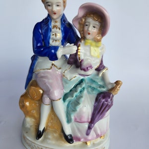 May include: A porcelain figurine of a couple in 18th-century attire. The man is wearing a blue jacket with gold trim and the woman is wearing a pink dress with a purple umbrella.