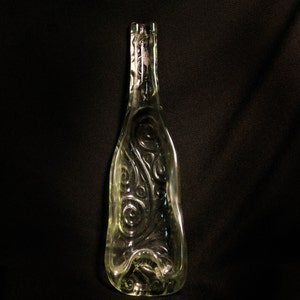 May include: Clear glass bottle with an elongated shape and decorative swirl patterns. The bottle has a narrow neck and a wide, rounded base. The glass has a slight green tint, and the bottle is set against a black background.