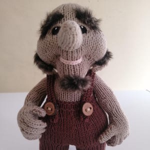 May include: A handmade knitted troll doll with a large nose, gray skin, and brown overalls. The doll has a whimsical design with faux fur accents around the face and a small pink collar. The doll is standing upright.