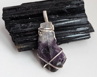 May include: A purple amethyst crystal pendant wrapped in silver wire. The pendant is displayed on a black stone.
