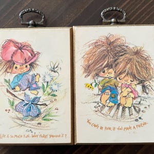 Op de afbeelding: Twee vintage ingelijste kunstwerken met illustraties van kinderen. Eén toont een kind met een roze hoed met de tekst "Life is so much fun... why hurry through it?". De andere toont twee kinderen met de tekst "You can't be poor if you have a friend."