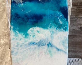 May include: A surfboard-shaped wall art piece featuring a glossy resin finish that depicts an ocean scene. The design transitions from a sandy tan base at the bottom to turquoise and deep blue water, topped with white lacy sea foam waves.