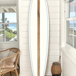 May include: A white surfboard with a central wooden stripe and a light blue outline. The surfboard is leaning against a white wall, suggesting a coastal or beach theme. The board's design is simple and elegant.