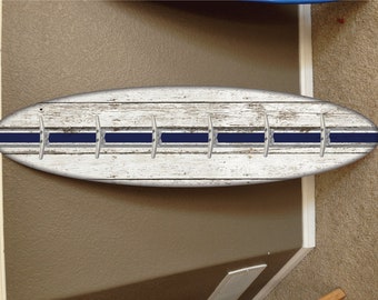 May include: Whitewashed wooden surfboard with a blue stripe and five silver hooks. The surfboard is mounted on a wall.