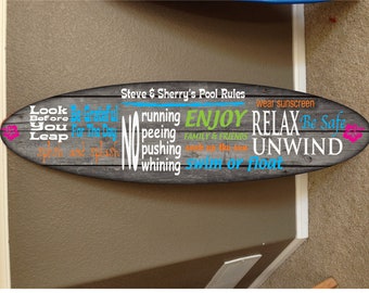 May include: A wooden surfboard sign with the text "Steve & Sherry's Pool Rules" and a list of pool rules, including "Look Before You Leap", "Be Grateful For The Day", "No Running", "No Peeing", "No Pushing", "No Whining", "Enjoy Family & Friends", "Relax Unwind", "Be Safe", "Swim or Float", and "Wear Sunscreen".