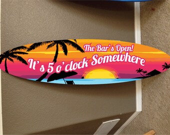May include: A colorful oval-shaped sign with a sunset scene and palm trees. The text reads "It's 5 o'clock Somewhere The Bar's Open!"