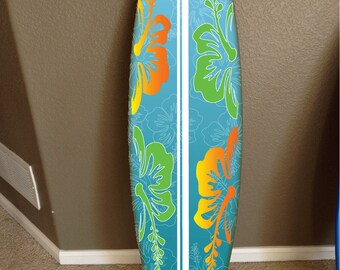 Op de afbeelding: Een blauw surfboard met een witte streep in het midden en een bloemenontwerp met gele, oranje en groene hibiscusbloemen.