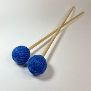 May include: Two blue yarn-wrapped mallet heads with natural wood handles. The mallets are angled across a white background. The blue yarn is tightly wound around the heads. The wooden handles are smooth and cylindrical.