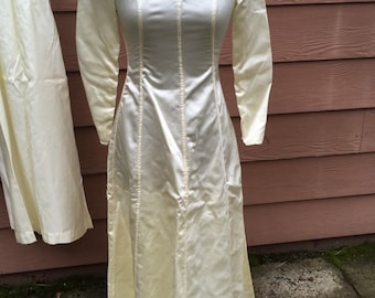 May include: A long, ivory satin dress with a round neckline and long sleeves. The dress has vertical seams down the front and back.
