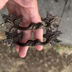 May include: Two matching drawer pulls made of dark bronze-toned metal. Each handle features an ornate, symmetrical design with intricate leaf-like scrollwork extending from the center grip outward to the decorative ends on both sides.