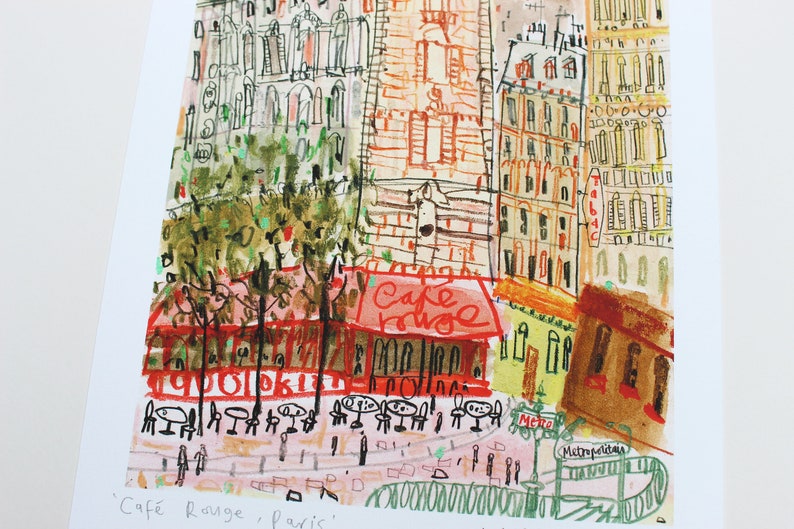 May include: A colorful illustration of a Parisian street scene with a cafe called "Cafe Rouge" and a sign that says "Metro" and "Metropolitan". The illustration is done in a whimsical style with a mix of watercolor and pen.