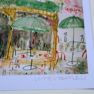 May include: Watercolor print of a Parisian cafe scene with green umbrellas, tables, and chairs. The print features the text "Deux Magots" and "Galeries". The artwork has a loose, expressive style with a focus on color and atmosphere.