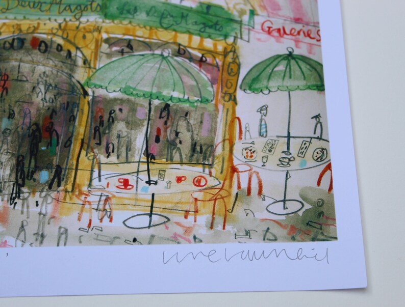 May include: Watercolor print of a Parisian cafe scene with green umbrellas, tables, and chairs. The print features the text "Deux Magots" and "Galeries". The artwork has a loose, expressive style with a focus on color and atmosphere.