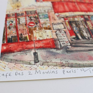 May include: Watercolor print of a Parisian cafe, featuring a red awning and outdoor seating. The artwork depicts the "Caf&eacute; Des 2 Moulins Paris" with the edition number 12/150. The image captures the essence of a charming European street scene.