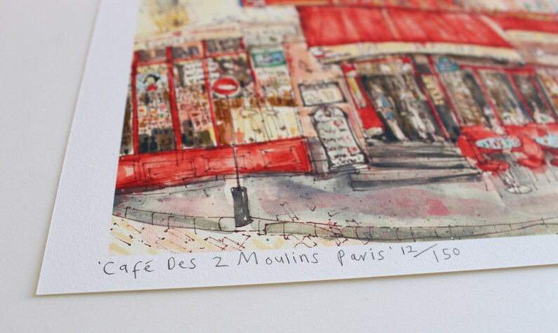 May include: Watercolor print of a Parisian cafe, featuring a red awning and outdoor seating. The artwork depicts the "Caf&eacute; Des 2 Moulins Paris" with the edition number 12/150. The image captures the essence of a charming European street scene.