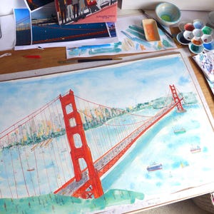 May include: A watercolor painting of the Golden Gate Bridge in vibrant orange-red, spanning across a light blue bay. The artwork features detailed suspension cables, a distant city skyline, and small ships on the water, rendered in a loose, artistic style.