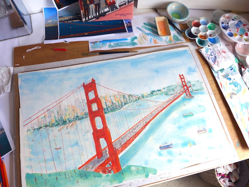 May include: A watercolor painting of the Golden Gate Bridge in vibrant orange-red, spanning across a light blue bay. The artwork features detailed suspension cables, a distant city skyline, and small ships on the water, rendered in a loose, artistic style.