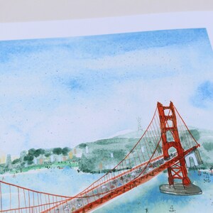 May include: A watercolor illustration of the Golden Gate Bridge in San Francisco, featuring a vibrant red suspension bridge spanning across blue water with green hills in the background and a light blue sky with soft, painted clouds above.