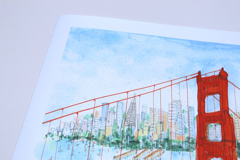 May include: A watercolor illustration of the Golden Gate Bridge in vibrant red, featuring thin suspension cables and a detailed city skyline in the background with blue water and small boats, all set against a light blue sky on white paper.