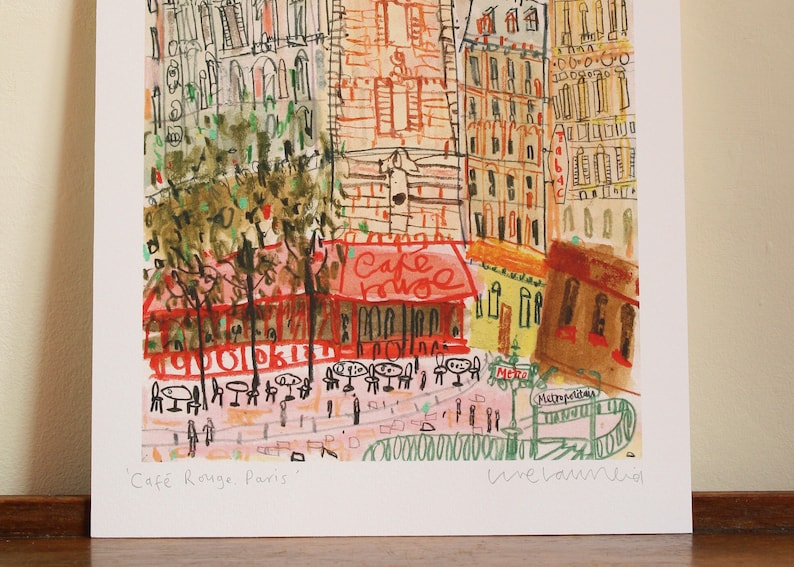May include: A watercolor painting of a Parisian street scene with a cafe called "Cafe Rouge" in the center. The cafe has red awnings and tables with chairs outside. The buildings are painted in a variety of colors, including yellow, orange, and brown. The painting is signed "Une Larme" in the bottom right corner.