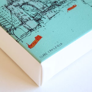 May include: A close-up of a turquoise canvas print with a black and white illustration of a city skyline. The print is framed with a white border and the artist's name, "CLARE CAULFIELD", is printed in black text on the bottom right corner.