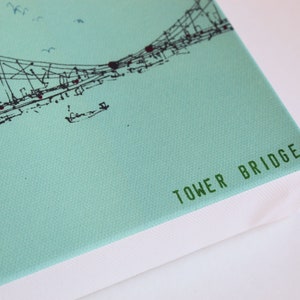 May include: A light blue canvas print with a black and white line drawing of the Tower Bridge. The bottom edge of the canvas is white and has the text "TOWER BRIDGE" printed in green.