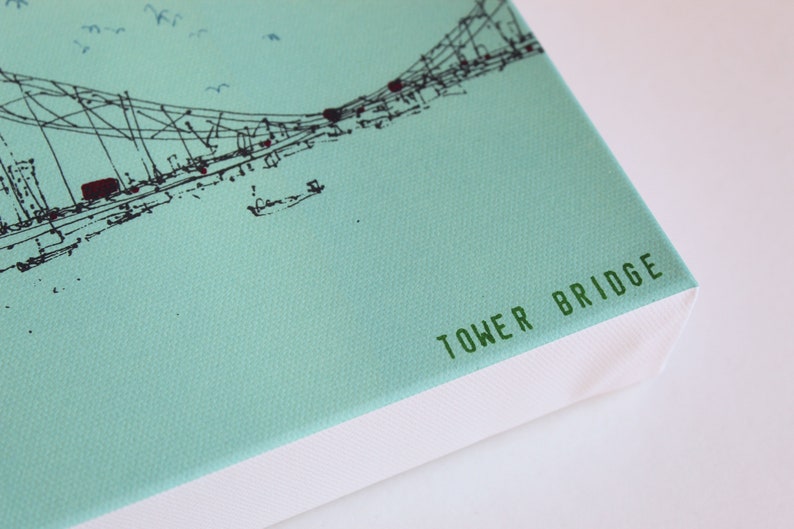May include: A light blue canvas print with a black and white line drawing of the Tower Bridge. The bottom edge of the canvas is white and has the text "TOWER BRIDGE" printed in green.