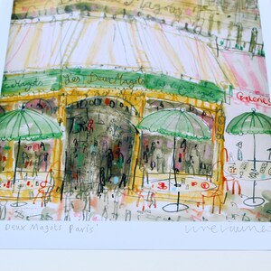 May include: Watercolor print of a Parisian cafe, "Les Deux Magots," with green umbrellas, outdoor seating, and the cafe's name in script. The artwork has a loose, expressive style with warm colors.