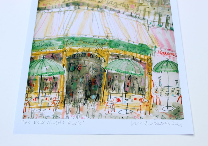 May include: Watercolor print of a Parisian cafe, "Les Deux Magots," with green umbrellas, outdoor seating, and the cafe's name in script. The artwork has a loose, expressive style with warm colors.