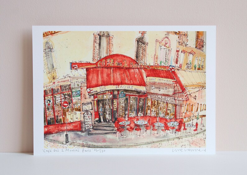 May include: Watercolor print of the Cafe des 2 Moulins in Paris, featuring a red awning and outdoor seating. The artwork depicts a street scene with buildings and the cafe's name visible. The print is numbered 15/150.