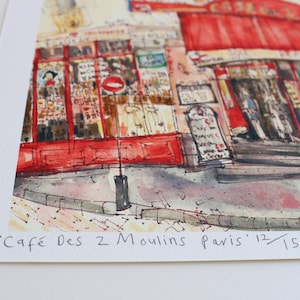 May include: Watercolor print of a Parisian cafe, featuring a red awning and outdoor seating. The artwork depicts the "Caf&eacute; Des 2 Moulins Paris" with the edition number 12/150. The street scene includes architectural details and a sidewalk.