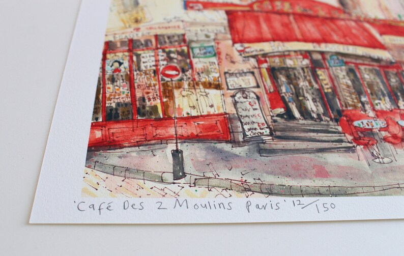 May include: Watercolor print of a Parisian cafe, featuring a red awning and outdoor seating. The artwork depicts the "Caf&eacute; Des 2 Moulins Paris" with the edition number 12/150. The street scene includes architectural details and a sidewalk.