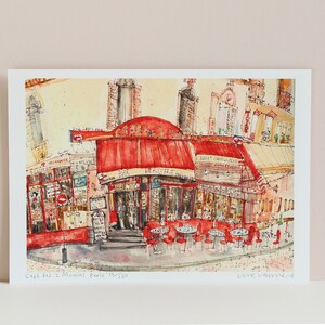 May include: Watercolor print of a Parisian cafe, featuring a red awning and outdoor seating. The artwork depicts the "Cafe des 2 Moulins Paris" with detailed architectural elements and street scenes. The print is on white paper.