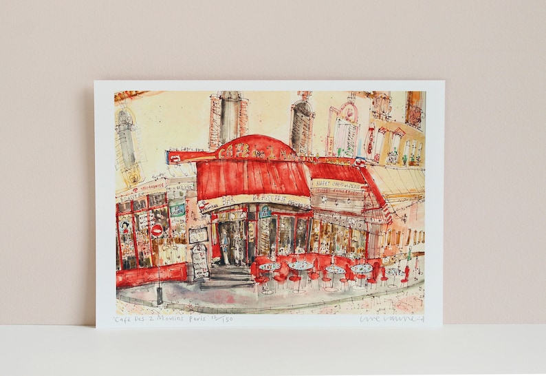May include: Watercolor print of a Parisian cafe, featuring a red awning and outdoor seating. The artwork depicts the "Cafe des 2 Moulins Paris" with detailed architectural elements and street scenes. The print is on white paper.