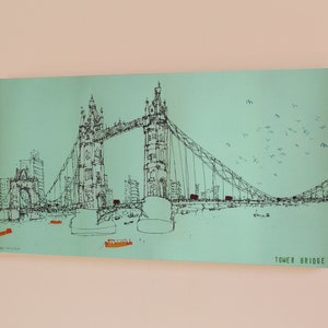 May include: A black and white line drawing of Tower Bridge in London, England. The bridge is depicted against a light blue background with a flock of birds flying overhead. The text "TOWER BRIDGE" is printed in black at the bottom right corner of the image.