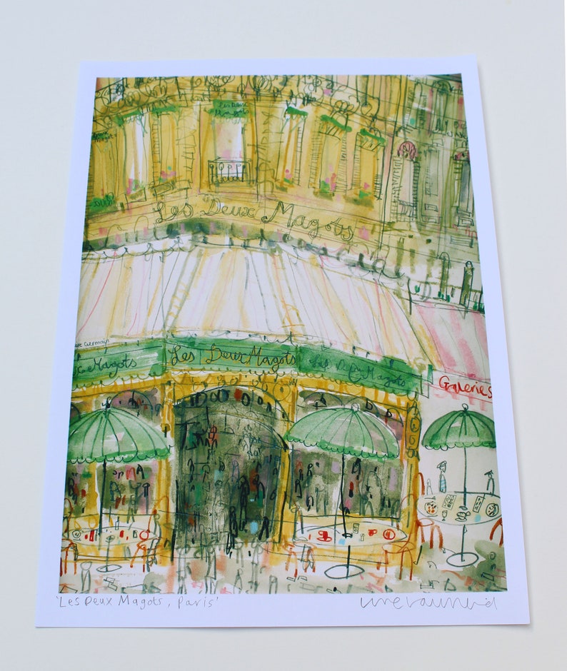 May include: Watercolor print of a Parisian cafe, "Les Deux Magots," with green awnings and umbrellas. The artwork features a detailed depiction of the building's facade, outdoor seating, and the cafe's name in cursive.