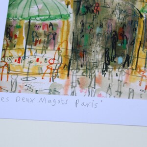 May include: An artistic print of a Parisian cafe scene, featuring green umbrellas, tables, and figures. The artwork has a loose, expressive style with a palette of yellows, greens, and reds. The text "Les Deux Magots Paris" is at the bottom.