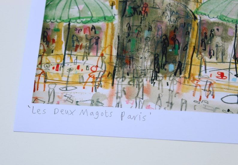 May include: An artistic print of a Parisian cafe scene, featuring green umbrellas, tables, and figures. The artwork has a loose, expressive style with a palette of yellows, greens, and reds. The text "Les Deux Magots Paris" is at the bottom.