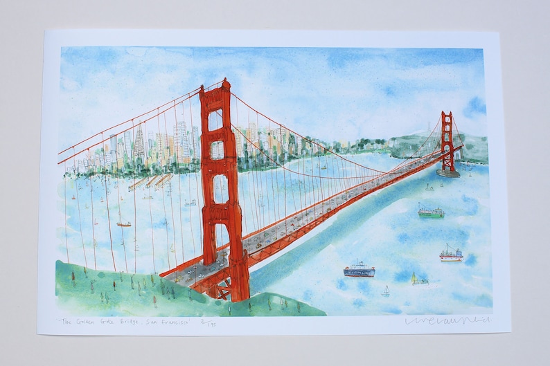May include: A watercolor print depicting the Golden Gate Bridge in San Francisco, featuring the iconic orange-red bridge spanning blue water with boats, a city skyline in the background, and the handwritten text 'The Golden Gate Bridge, San Francisco'.