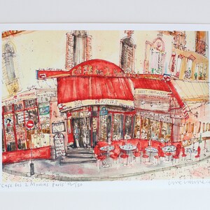 May include: Watercolor print of a Parisian cafe, "Cafe des 2 Moulins", with a red awning and outdoor seating. The artwork features detailed architectural elements and street scenes, capturing the essence of a French bistro.