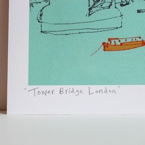 May include: A turquoise blue print of a stylized drawing of Tower Bridge in London. The print includes a small boat in the foreground and the text 'Tower Bridge London'.