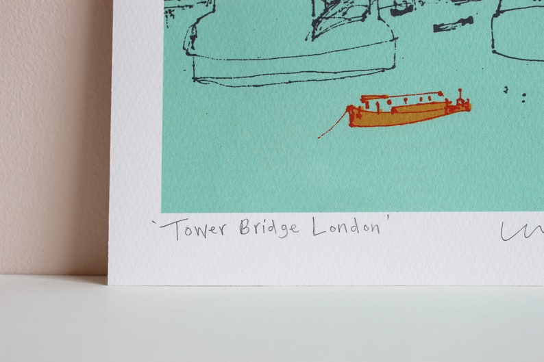 May include: A turquoise blue print of a stylized drawing of Tower Bridge in London. The print includes a small boat in the foreground and the text 'Tower Bridge London'.