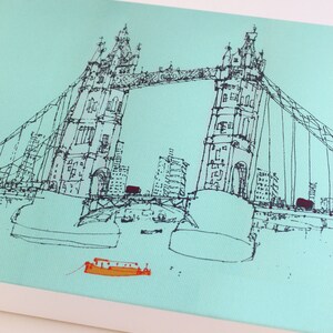 May include: A black and white line drawing of the Tower Bridge in London, England. The bridge is drawn in a simple, minimalist style, with the two towers and the suspension bridge clearly visible. There are small boats in the water below the bridge.