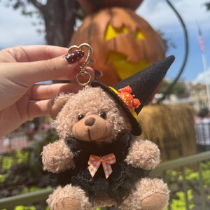 May include: A plush teddy bear keychain featuring a tan fuzzy body, a black witch hat adorned with a small orange pumpkin, and a black lace dress accented with a small orange and white checkered bow, attached to a gold heart-shaped metal clasp.