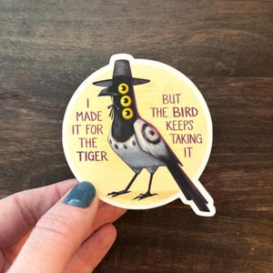 May include: A sticker featuring a stylized bird wearing a hat with three yellow eyes. The bird is black, white, and gray, set against a yellow circle. Text on the sticker reads "I made it for the tiger but the bird keeps taking it."