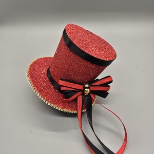 May include: A miniature red glitter top hat with a black band and a gold-trimmed brim. The hat is adorned with a black and red bow and gold beads, with black and red ribbons hanging down. This decorative hat is suitable for themed parties.