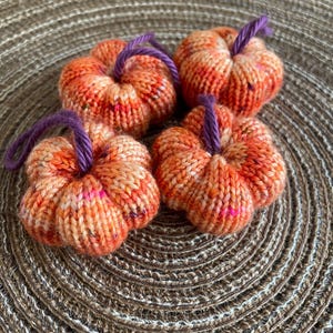 May include: Four small, hand-knitted pumpkins made from variegated orange yarn with speckled accents. Each pumpkin features a short, dark purple yarn stem looped at the top. The pumpkins have a rounded, ribbed shape and a soft, textured appearance.